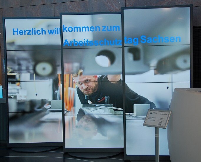 Drei Videostelen stehen nebeneinander. Darauf ist ein Bild mit einem Arbeiter in schwarzer Arbeitskleidung zu sehen. Er kontrolliert die technische Einstellung an der Werkzeugmaschine