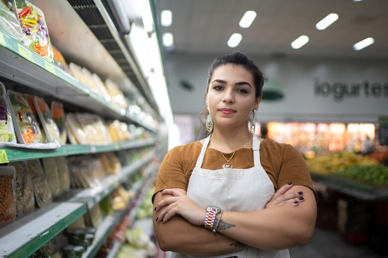 Junge Frau mit weißer Schürze steht mit verschränkten Armen vor einem Verkaufsregal